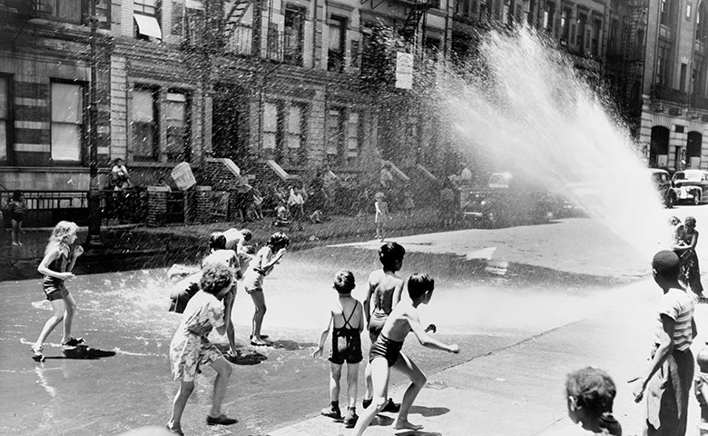 A black and white photo of children, some wearing bathing suits play in the street with a fire hydrant spraying water.