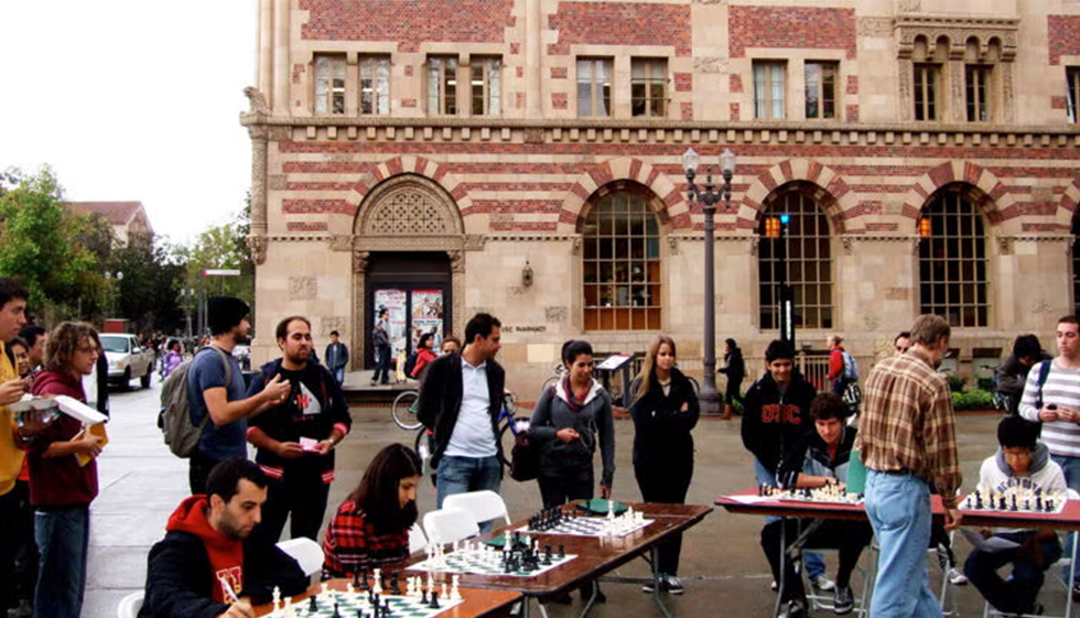 A man in a plaid shirt plays multiple chess games simultaneously at outdoor tables, while a crowd watches and participates in a university courtyard.