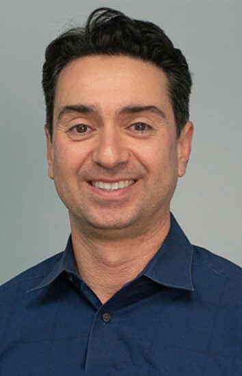 Headshot of Kamy Akhavan smiling and wearing a dark blue, collared, button-down shirt.
