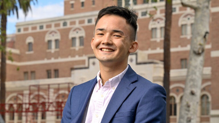 A young man, Liam Tsao, smiles while wearing a blue sport coat over a light pink dress shirt and stands in front of a large, brick USC library building and trees.