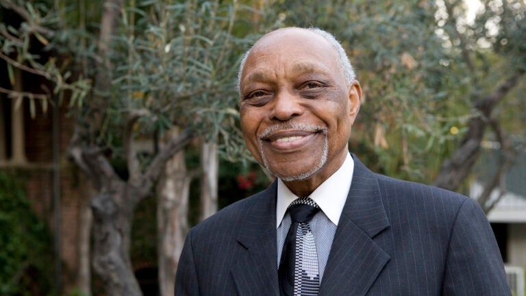 The Rev. Dr. Cecil L. Murray smiles at the camera wearing a suit and tie and standing in front of a background of trees