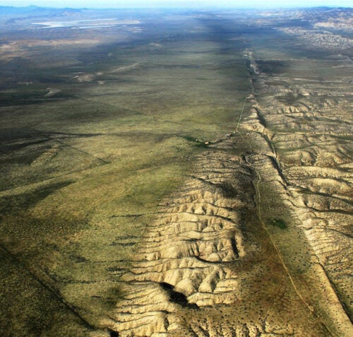 An aerial view of a vast desert landscape showing barren terrain with patterns of dry washes and sparse vegetation. The terrain is uneven with areas of slight elevation changes giving it a wrinkled texture.