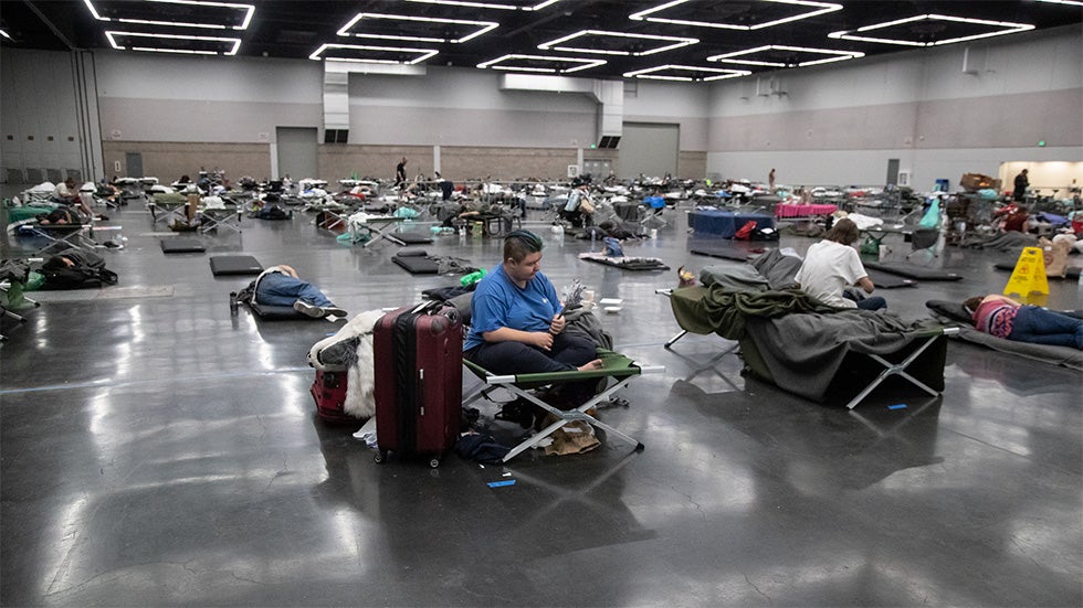 A large shelter with rows of cots, people resting and sitting, and personal belongings scattered around.