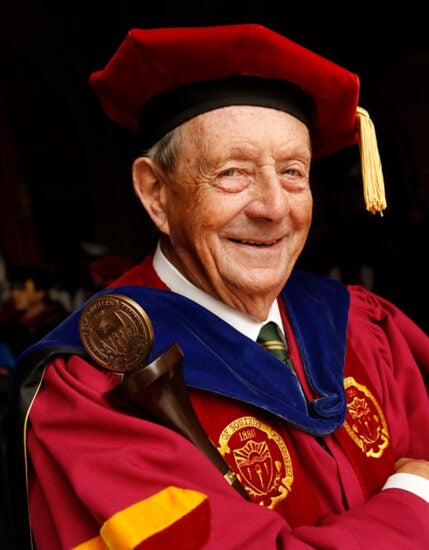 John Callaghan poses with arms crossed, smiling and leaning toward the camera in full university marshal regalia