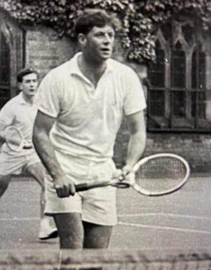 Black and white image of young John Callaghan wearing white tennis attire holding a racket at the ready while playing doubles tennis