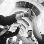 A black and white photo shows a group of people inside a church, holding hands and raising them together in a gesture of unity.