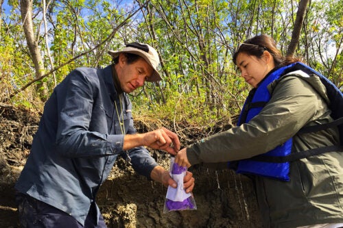 Two researchers collect soil samples from a riverbank, using a small bag to store the samples.