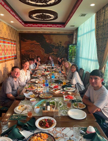 Students sit around a long table in a narrow room with an array of foods dispersed on the table