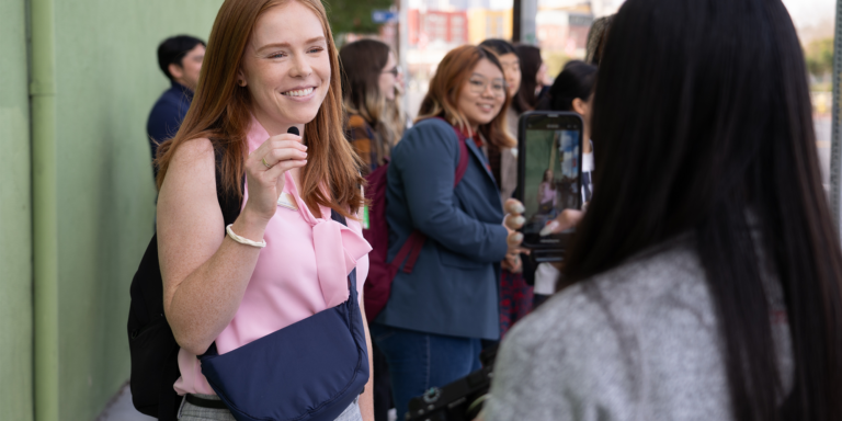 A woman smiling while speaking into a microphone, surrounded by a group of people, with one person recording her on a smartphone.