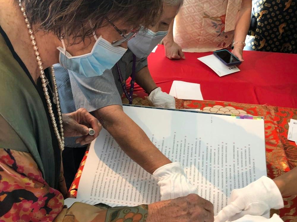 A person's hands hold a small stamp while other, gloved, hands point to a specific area on a page in a large book.