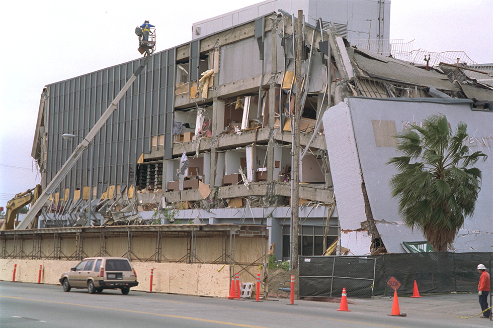 A man in a boom lift surveys a heavily damaged building as a car passes below