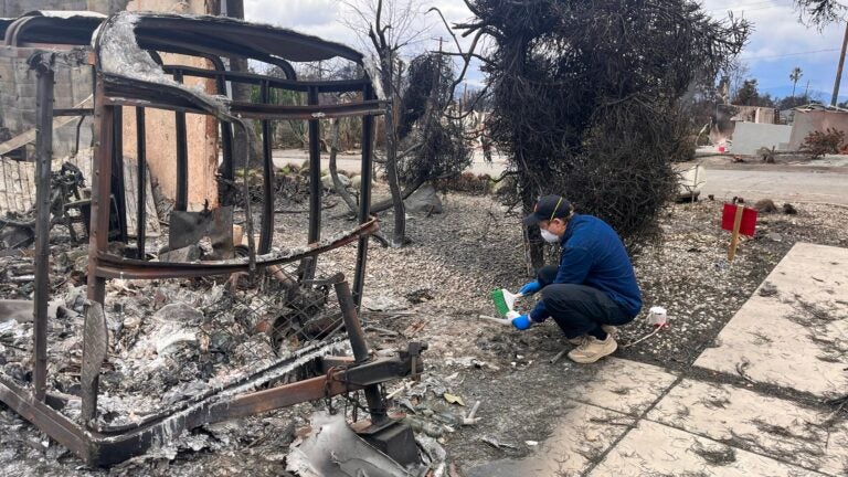 Masked person collecting soil near the charred remains of a home