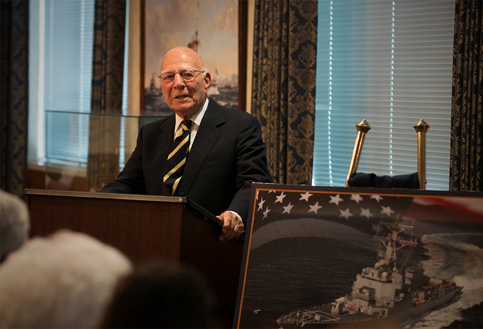 Paul Ignatius speaks from behind a lectern near an artist’s depiction of the USS Paul Ignatius