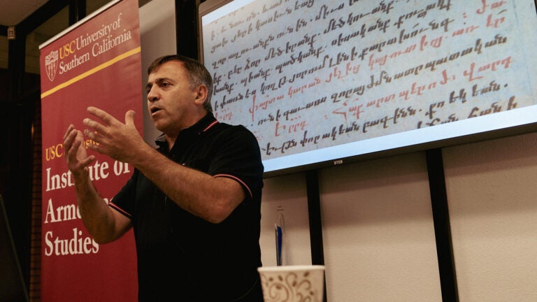 Linguist Hrach Martirosyan stands at a podium in front of a whiteboard displaying Armenian writing