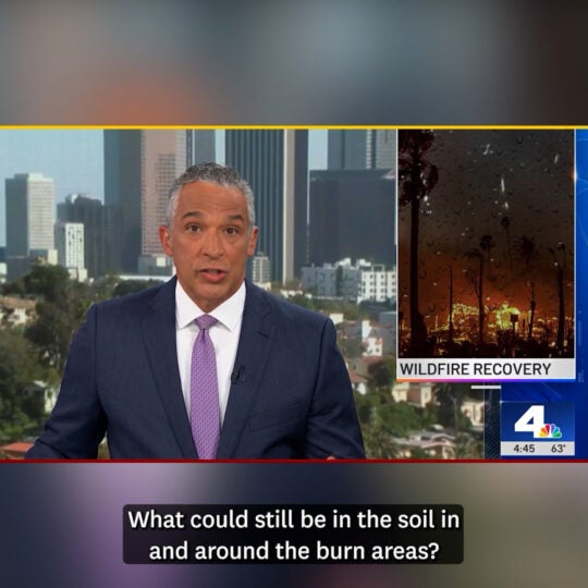News anchor sits at desk with video images behind him