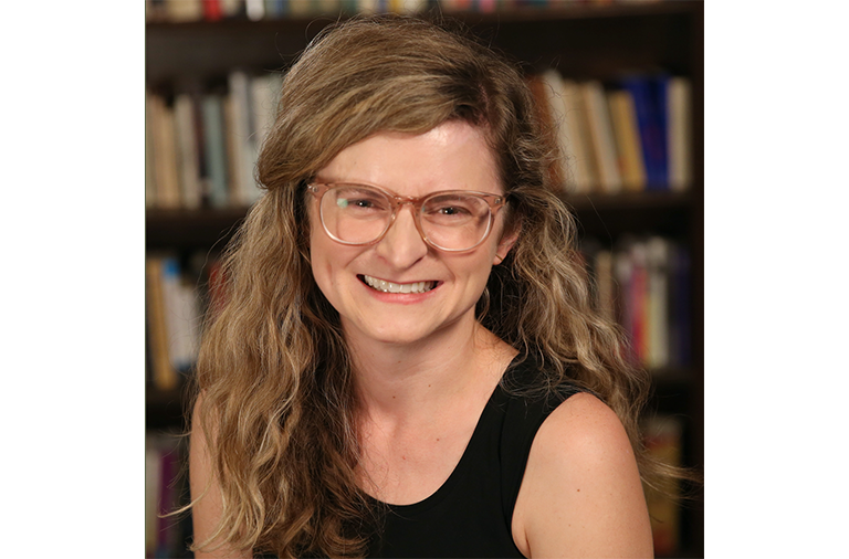 Amy, who is wearing rose-colored clear glasses and a black top, smiles in front of bookcases.