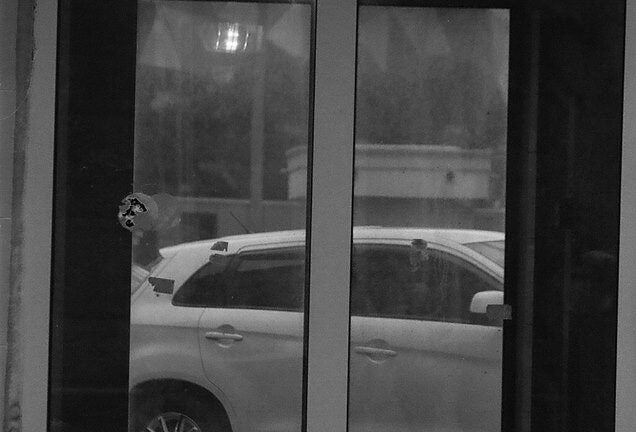 Black and white photo of white car, as seen through reflection of glass