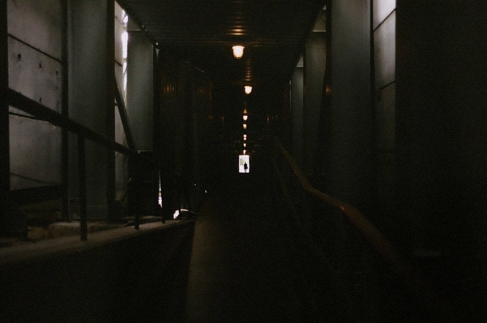 Photo of dark tunnel(?)/walkway with overhead lighting. At the end of the walkway is light and a person's silhouette.