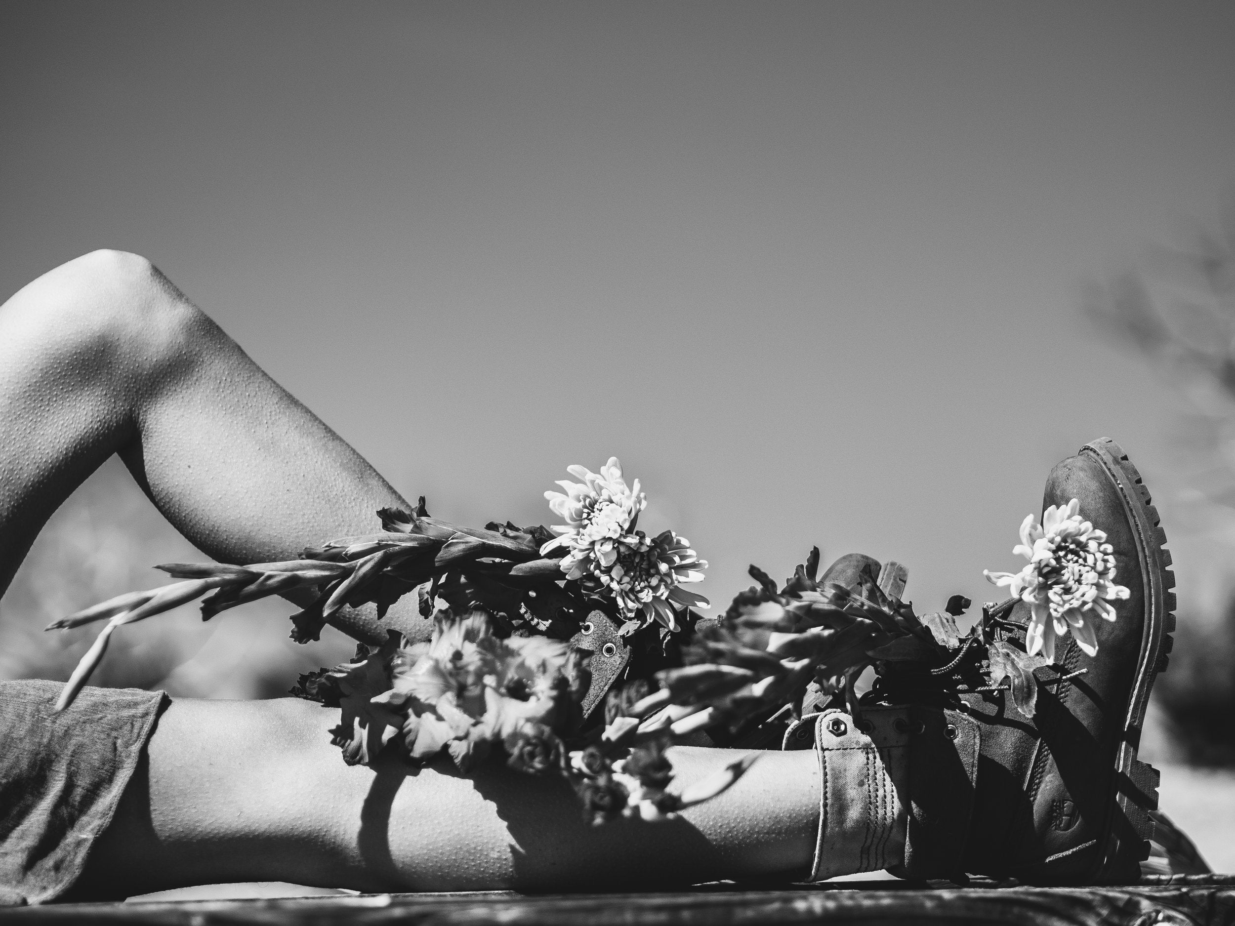 Black-and-white photo of a person's legs as they lie down. They are wearing boots and have flowers placed on their legs.