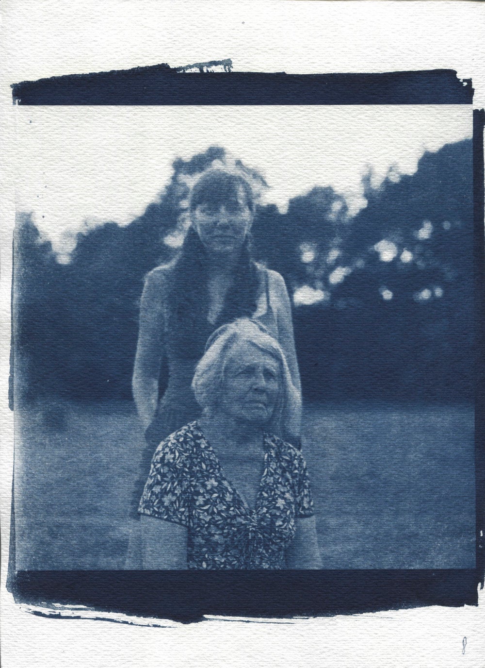 Two older women, one standing, one sitting, in grass with trees in the background