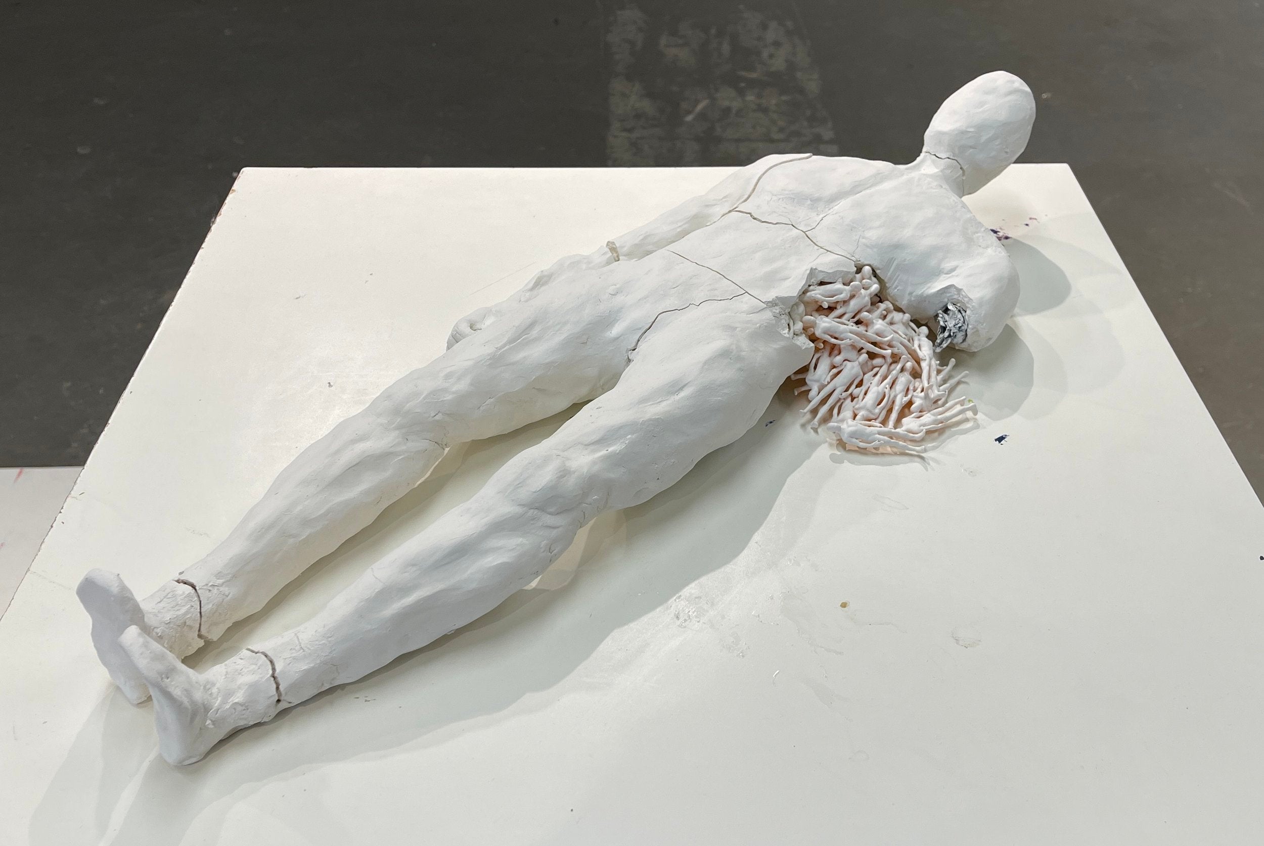 Sculpture of person lying down with a fracture on their side. From the fracture emerges smaller replicas of the larger sculpture.