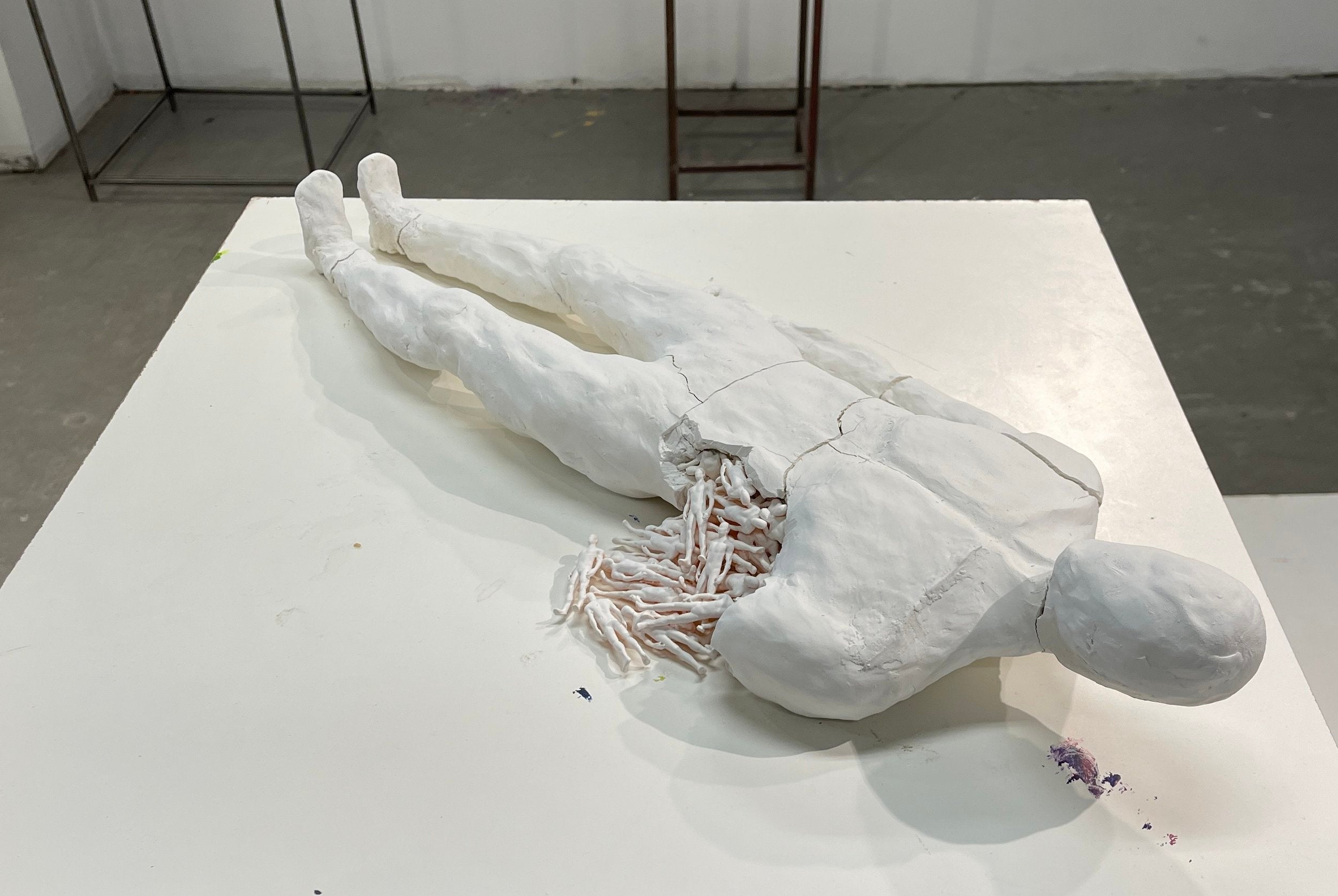 Sculpture of person lying down with a fracture on their side. From the fracture emerges smaller replicas of the larger sculpture.