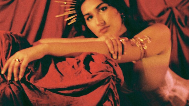 A young woman wearing a gold sun headpiece poses in front of a draped red curtain