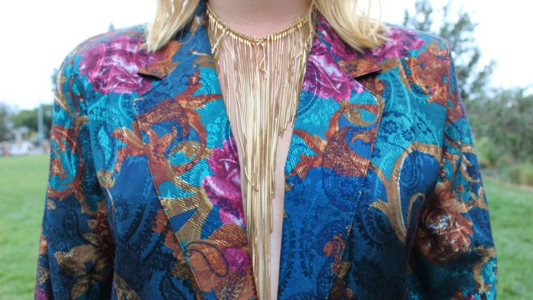 A woman with blonde hair wears a gold necklace and decorative blazer.