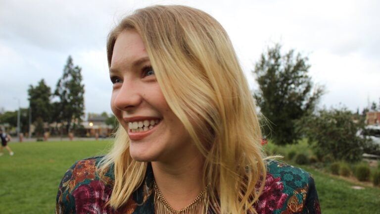 A woman with blonde hair smiles as she wears a gold necklace and decorative blazer.