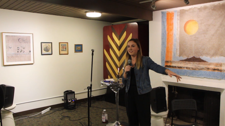 Kyra Brown doing stand up set, holding a microphone, in a room with framed wall art.