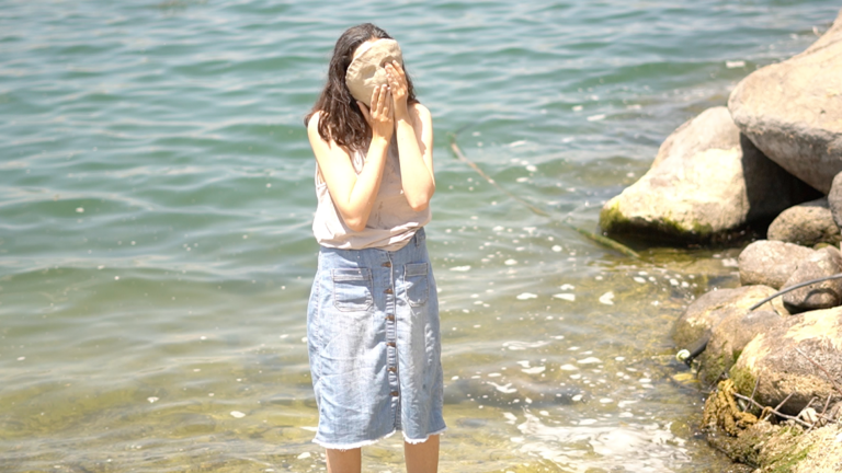 Broome standing in the ocean by some rocks, holding her mask sculpture to her face.