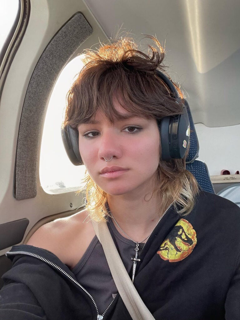 Person facing the camera, wearing large over-ear headphones, picture taken as a selfie inside a vehicle, with seatbelt and rounded window visible, wearing a Jurassic Park sweatshirt, falling off one shoulder, shaggy layered hair, septum piercing and dagger necklace visible.