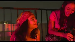 Two girls with medium length brown hair, one with a bandana on, smiling on a balcony at night, lit redly