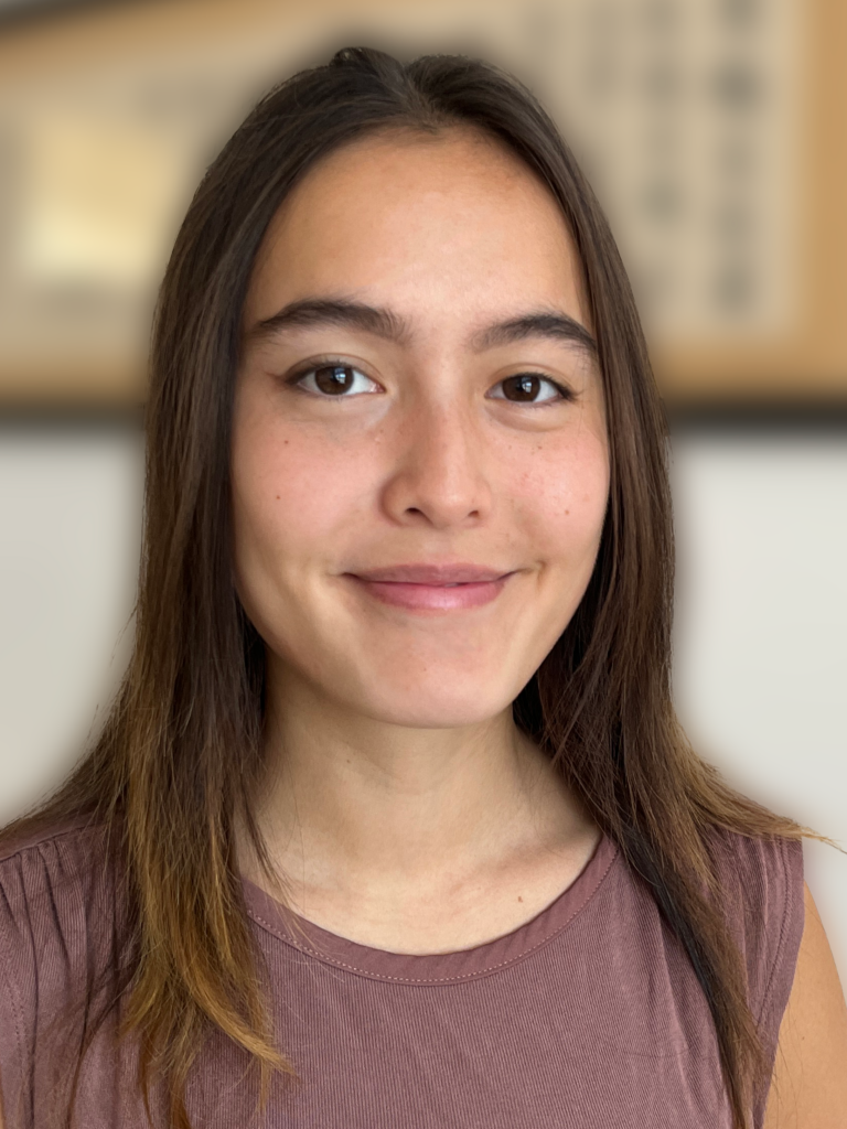 Woman stands in front of a blurred indoor background and smiles with closed mouth.