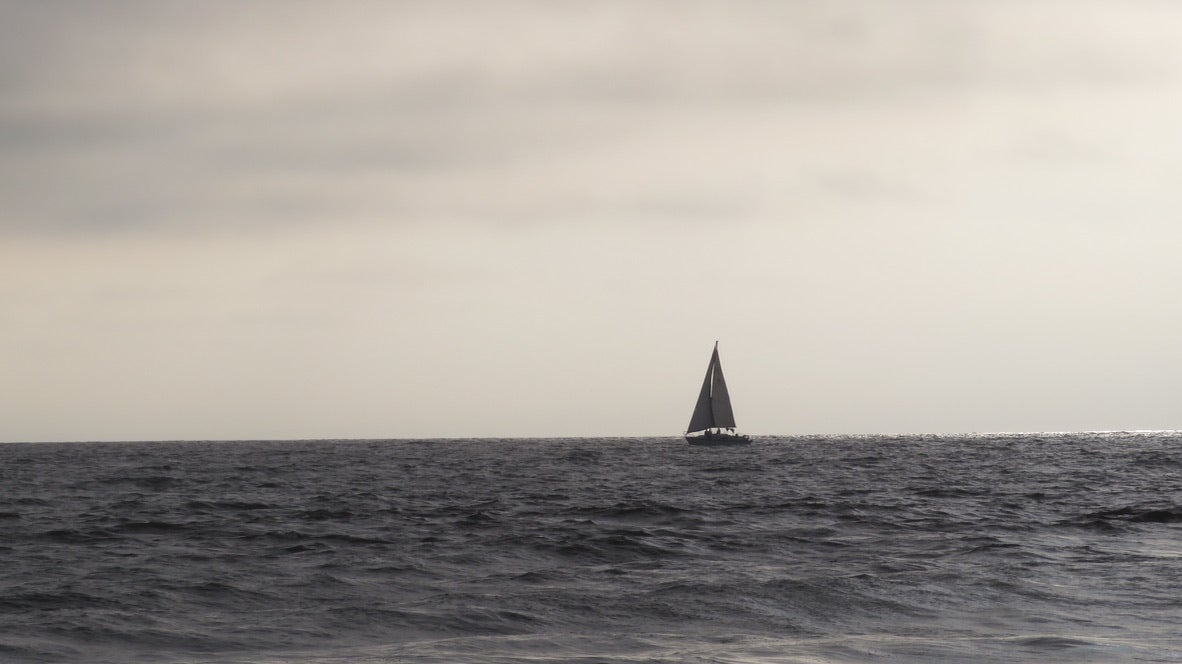 The silhouette of a sailboat on the horizon of a dark gray ocean and cloudy gray sky.