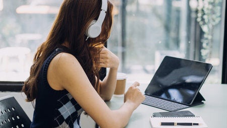 A woman with long dark hair has a white headphone on, and is working on her computer.
