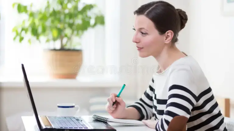 Woman is taking notes in her notebook, staring into her laptop.