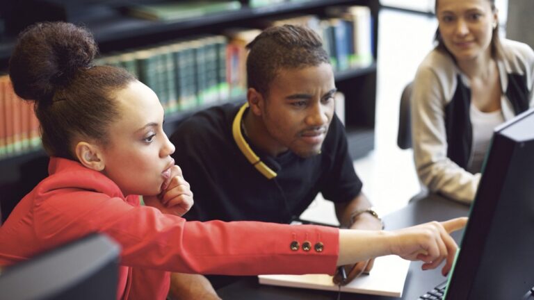 3 students, 2 women and 1 male is seated together, one woman is pointing at the screen off to the side and the guy is seated in the middle and both him and the other woman is looking into the laptop screen that their fellow classmate is pointing to.