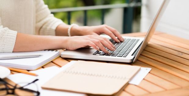 Cropped photo of a female typing at her laptop, on her desk are papers and her tablet