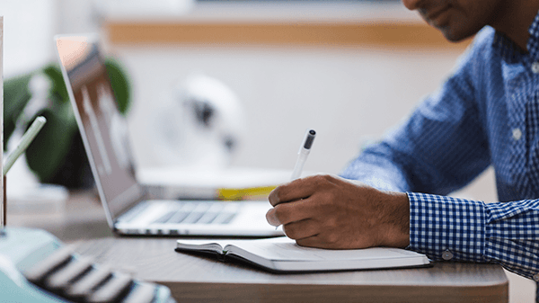 Black male student writing down on paper while looking at his laptop