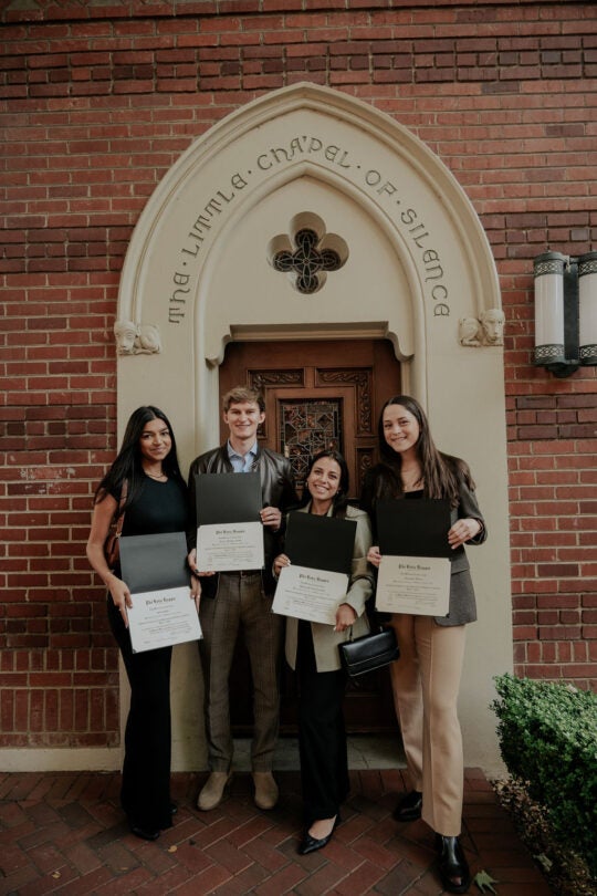 Four Phi Beta Kappa Inductees standing in front of doorway to Little Chapel of Silence, holding certificates and smiling.