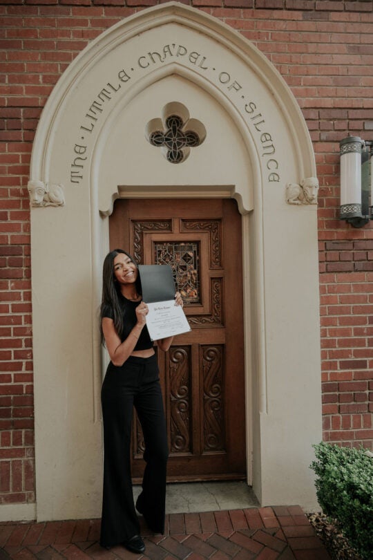 Inductee standing at the doorway to the Little Chapel of Silence, holding membership certificate up above chest, and smiling.