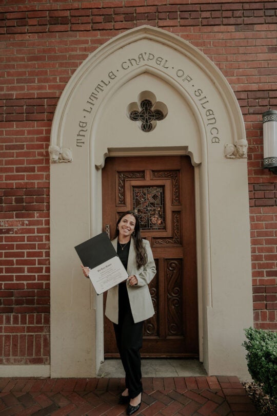 Inductee smiling and holding membership certificate, standing in front of doorway to the Little Chapel of Silence.
