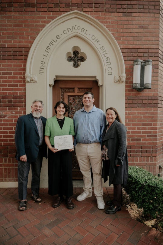Inductee smiling and holding membership certificate, standing in front of doorway to the Little Chapel of Silence with three guests.