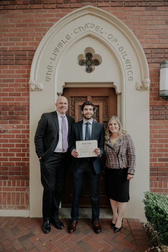 Inductee smiling and holding membership certificate, standing in front of doorway to the Little Chapel of Silence with two guests.
