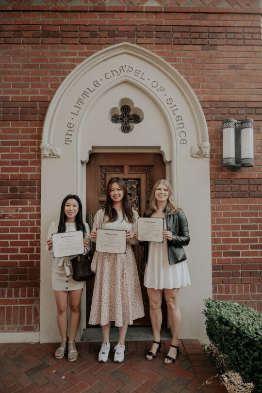 Three inductees smiling and holding membership certificate, standing in front of doorway to the Little Chapel of Silence.