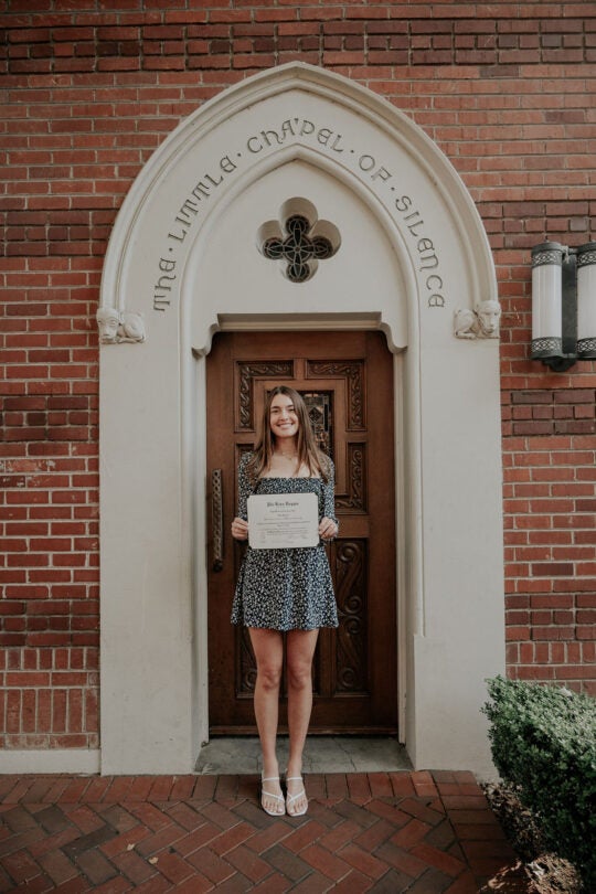 Inductee smiling and holding membership certificate, standing in front of doorway to the Little Chapel of Silence.