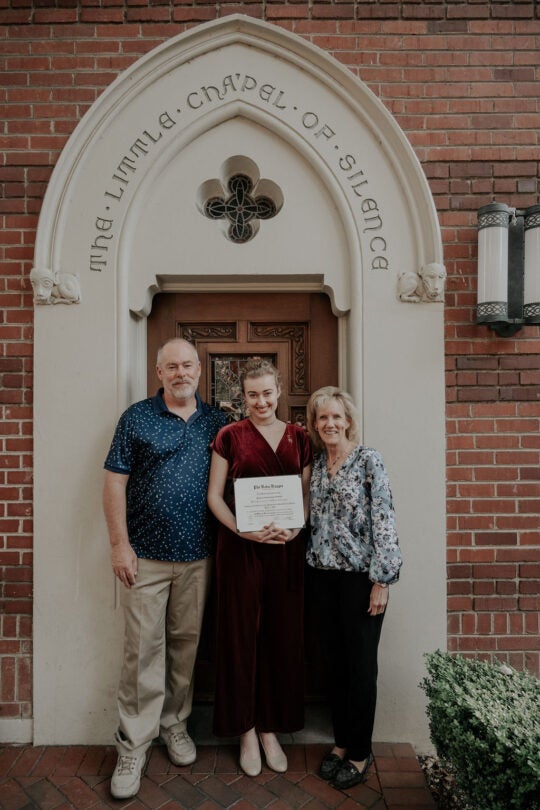 Inductee holding membership certificate, standing with two guests at doorway to the Little Chapel of Silence. All are smiling.