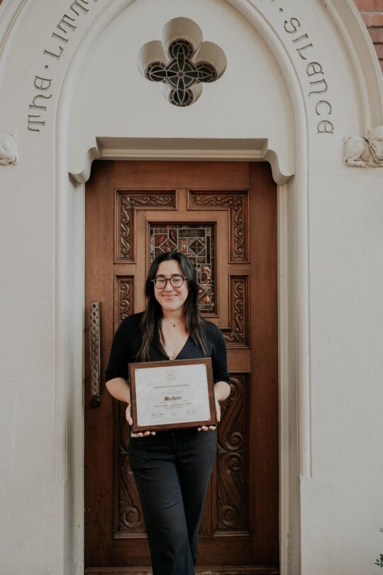 Award recipient Mia Speier standing at doorway to the Little Chapel of Silence, holding award plaque.