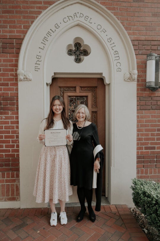 Inductee smiling and holding a membership certificate. Standing at doorway to the Little Chapel of Silence with a guest.